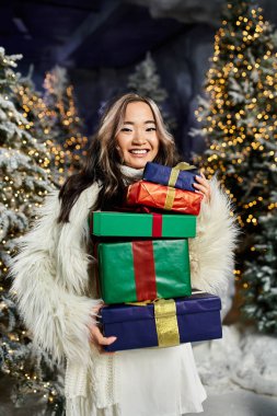 Genç bir kadın, muhteşem Noel süslemelerinin arasında hediyeler tutarken ışıl ışıl gülümsüyor..
