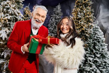 A cheerful man and woman exchange gifts while surrounded by snowy trees and holiday decorations. clipart