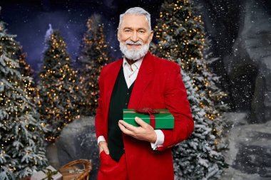 A smiling gentleman dressed in a vibrant red outfit presents a wrapped gift in a snowy setting. clipart