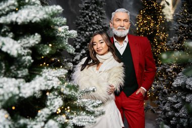 Bir erkek ve kadın şık kıyafetler içinde, parıldayan Noel ağaçlarıyla çevrili bir şekilde poz verirler..