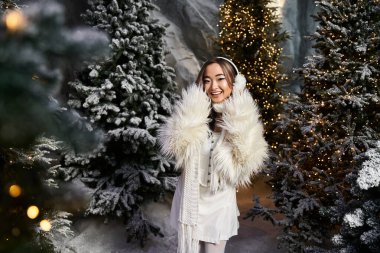 Etrafı güzel ışıklandırılmış Noel ağaçları ve karlarla çevriliyken genç bir kadın sıcacık gülümsüyor..