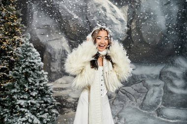 A young woman dressed in a beautiful winter outfit enjoys the festive snowfall surrounded by trees. clipart