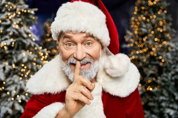 A jolly figure in red and white playfully asks for quiet amid twinkling holiday decorations.