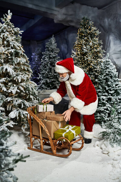 Санта Клаус аккуратно организовывает подарки на деревянных санях в окружении сверкающих деревьев.
