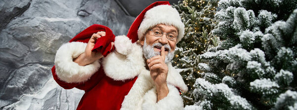 Радостная фигура в красном костюме делится секретом в окружении праздничной зелени и снега.