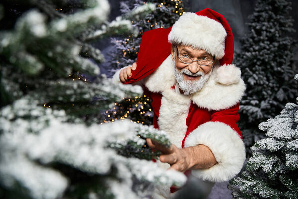 Праздничная фигура в красно-белом цвете протягивает руку среди заснеженных рождественских елок, распространяя радость.