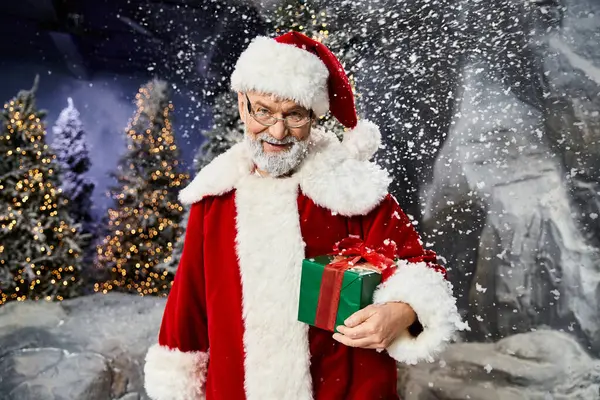 Noel Baba kostümü giymiş neşeli bir şahsiyet, kış harikalar diyarının ortasında paketlenmiş bir hediye sunar..