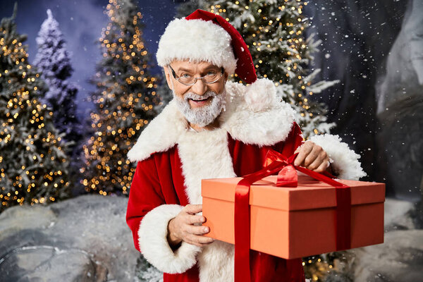 Санта Клаус с радостью представляет красочный подарок среди заснеженных деревьев и мерцающих огней.