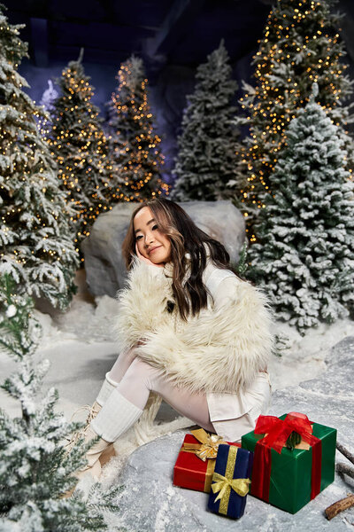 A young woman sits happily in a winter wonderland surrounded by festive trees and wrapped gifts.