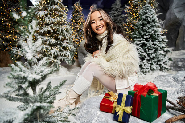 Радостная фигура, одетая в зимний наряд, представляет красочные подарки среди снежных деревьев.