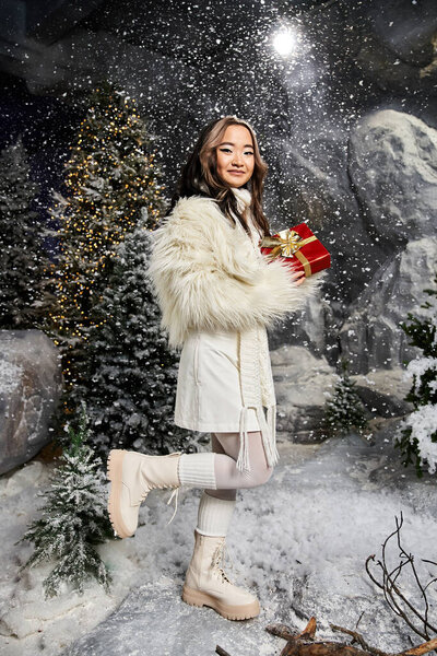 Женщина, одетая в зимний наряд, радостно представляет завернутый подарок среди снежных деревьев и украшений.