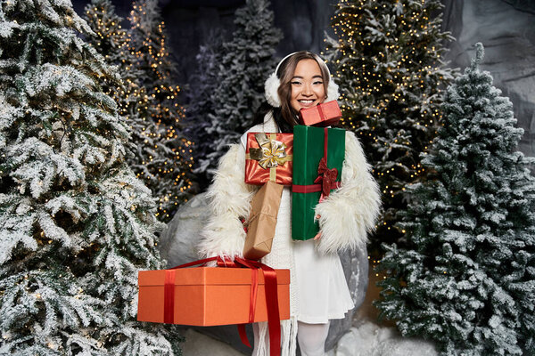 Веселая женщина в зимней одежде стоит среди снежных деревьев, держа радостные праздничные подарки.