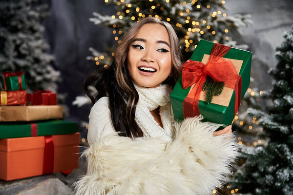 Восхитительная женщина демонстрирует красочный подарок среди праздничной рождественской обстановки с елками.