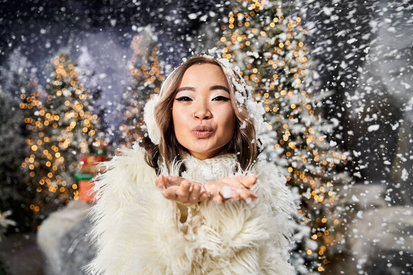 Человек в зимнем наряде выдувает снежинки в воздух, окруженный праздничными украшениями.