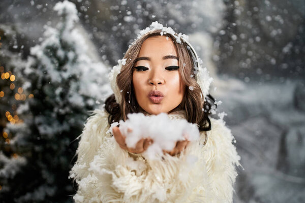 A young woman plays in a snowy landscape, blowing flurries of snow into the air while smiling.