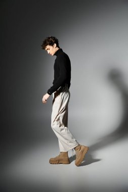 Zarif bir genç adam, siyah balıkçı yakasını ve açık renk pantolonunu bir stüdyo ortamında göstererek zarif bir şekilde ayakta duruyor..