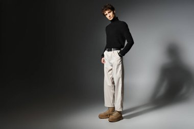 Kıvırcık saçlı yakışıklı genç bir adam dramatik koyu gri arka planda modern bir kıyafet sergiliyor..