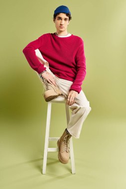 Pembe süveter ve bej pantolon giyen şık bir genç adam pastel bir arka fona karşı güvenli bir şekilde bir taburede oturur..