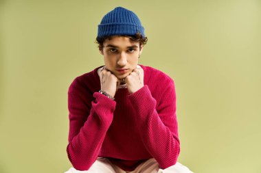 Canlı pembe süveter giyen yakışıklı genç bir adam, pastel yeşil arka planda, gündelik giyinmiş, dalgın bir şekilde oturur..