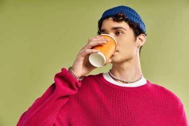 Yakışıklı genç bir adam turuncu bardaktan yudumluyor, rahat pembe bir kazak ve mavi bir bere giyiyor..