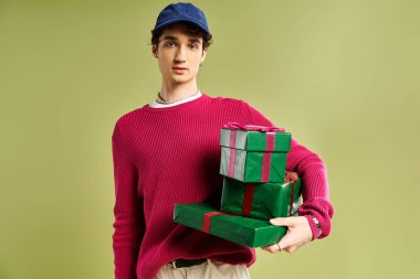Parlak kazaklı şık bir genç adam yeşil bir arkaplan karşısında güzel paketlenmiş Noel hediyelerini mutlulukla tutuyor..