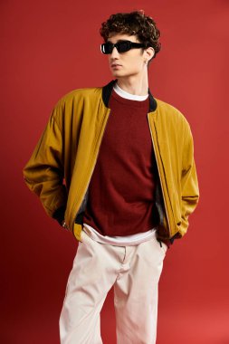 Kıvırcık saçlı yakışıklı genç bir adam sarı bir ceketle çarpıcı bir kırmızı zemine karşı güvenle poz veriyor..