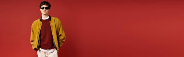 Kıvırcık saçlı, sarı ceketli genç bir adam dikkat çekici bir kırmızı arka plan sergiliyor..