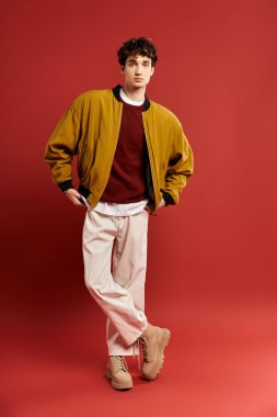 Kıvırcık saçlı şık bir genç adam, koyu kırmızı arka planda sarı bir ceketle rahatça poz verir..