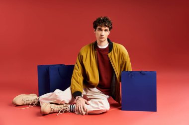 A handsome young man sits on the floor holding shopping bags while dressed in trendy attire during Black Friday. clipart