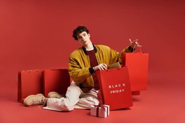 Şık bir genç adam Kara Cuma 'da renkli alışveriş torbalarıyla çevrili yerde oturuyor ve heyecanını gösteriyor..