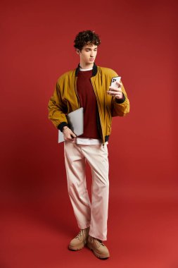 Kıvırcık saçlı yakışıklı genç bir adam elinde bir telefon ve tablet ile kırmızı üzerinde sarı bir ceketle poz veriyor..