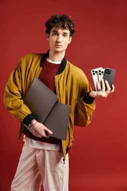 A stylish young man with curly hair holds tech devices while posing confidently on a bold red background. clipart