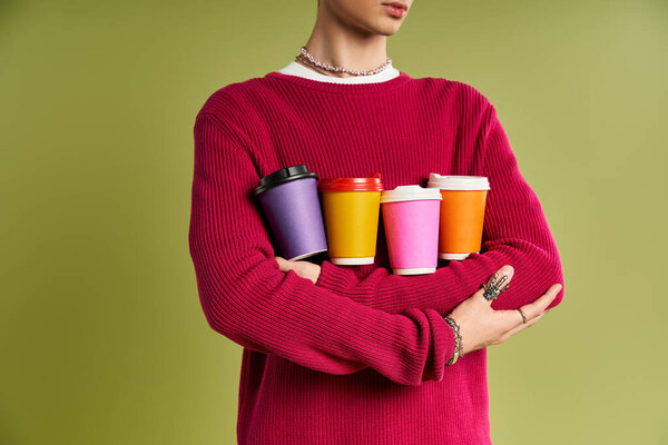 Красивый молодой человек в розовом свитере уверенно позирует с яркими кофейными чашками, демонстрируя свой уникальный стиль.