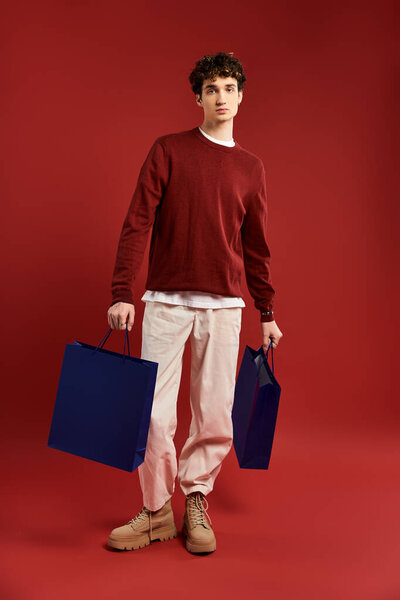 Красивый молодой человек с вьющимися волосами уверенно стоит, держа сумки после успешной Черной пятницы.