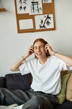 A young man relaxes on a couch, wearing headphones and smiling as he listens to music. clipart