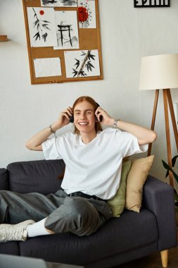 Neşeli bir yüzü olan genç bir adam kanepede rahatça oturur ve müzik dinler..