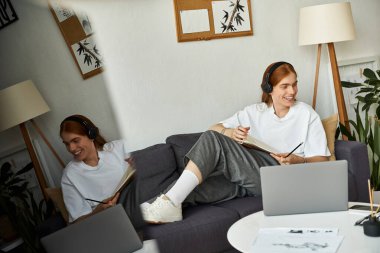 Genç bir adam kanepeye oturmuş, bitkilerle çevrili kulaklık takmış, mutlu bir şekilde çizim yapıyor..