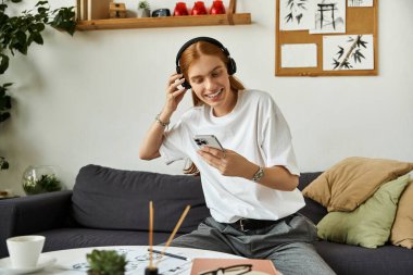 Genç bir adam neşeyle gülümser, kulaklıkla rahat bir ortamda müzik dinler..