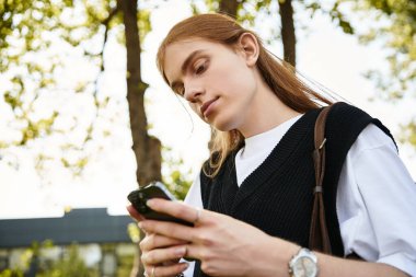 Uzun saçlı genç bir adam parktaki canlı yeşilliğin ortasındaki akıllı telefonuna odaklanıyor..