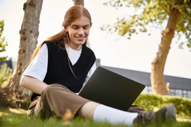 A young man sits on the grass, focused on his laptop, with a smile on his face. clipart