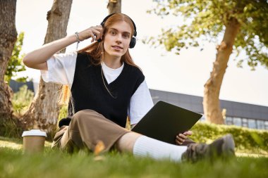Genç adam sakin bir parkta dizüstü bilgisayarıyla çalışırken müzik dinliyor..