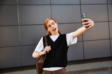 Genç adam, modern bir arkaplana karşı şık bir kıyafetle selfie çekerken gülümsüyor..