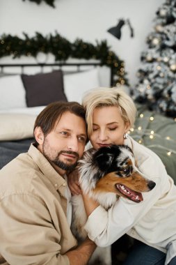 A couple enjoys a warm embrace with their dog, surrounded by holiday decorations at home. clipart