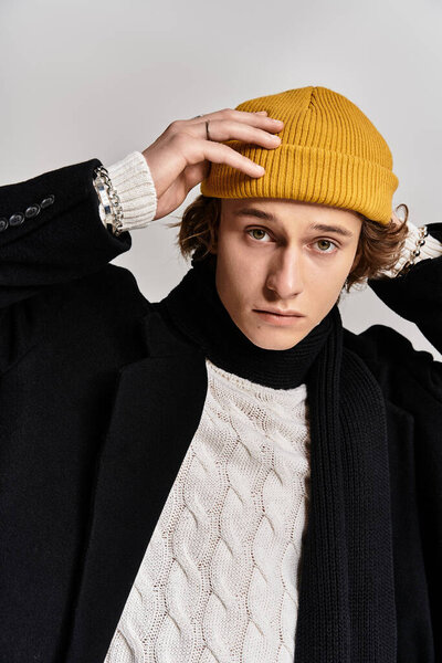 A handsome young man adjusts his yellow beanie while wearing a chic black coat and a cozy scarf.