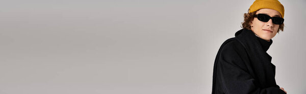 A stylish young man models a black coat and bright yellow beanie in a studio.