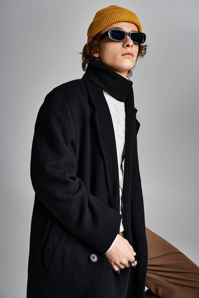 Красивый молодой человек стоит с уравновешенностью, одетый в черное пальто и желтую шапочку в гладкой студии.