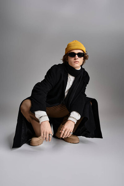 A dapper young man in a black coat and yellow beanie crouches elegantly, showcasing his unique style and confidence.