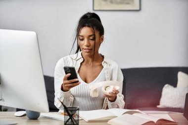 Genç ve güzel bir kadın evde çalışıyor, elinde kahve tutarken akıllı telefonuna odaklanıyor..