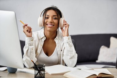 A cheerful young woman studies at home, wearing headphones and smiling while focused on her work. clipart