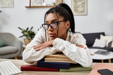 A focused woman rests her chin on books, deep in thought while studying online at home. clipart